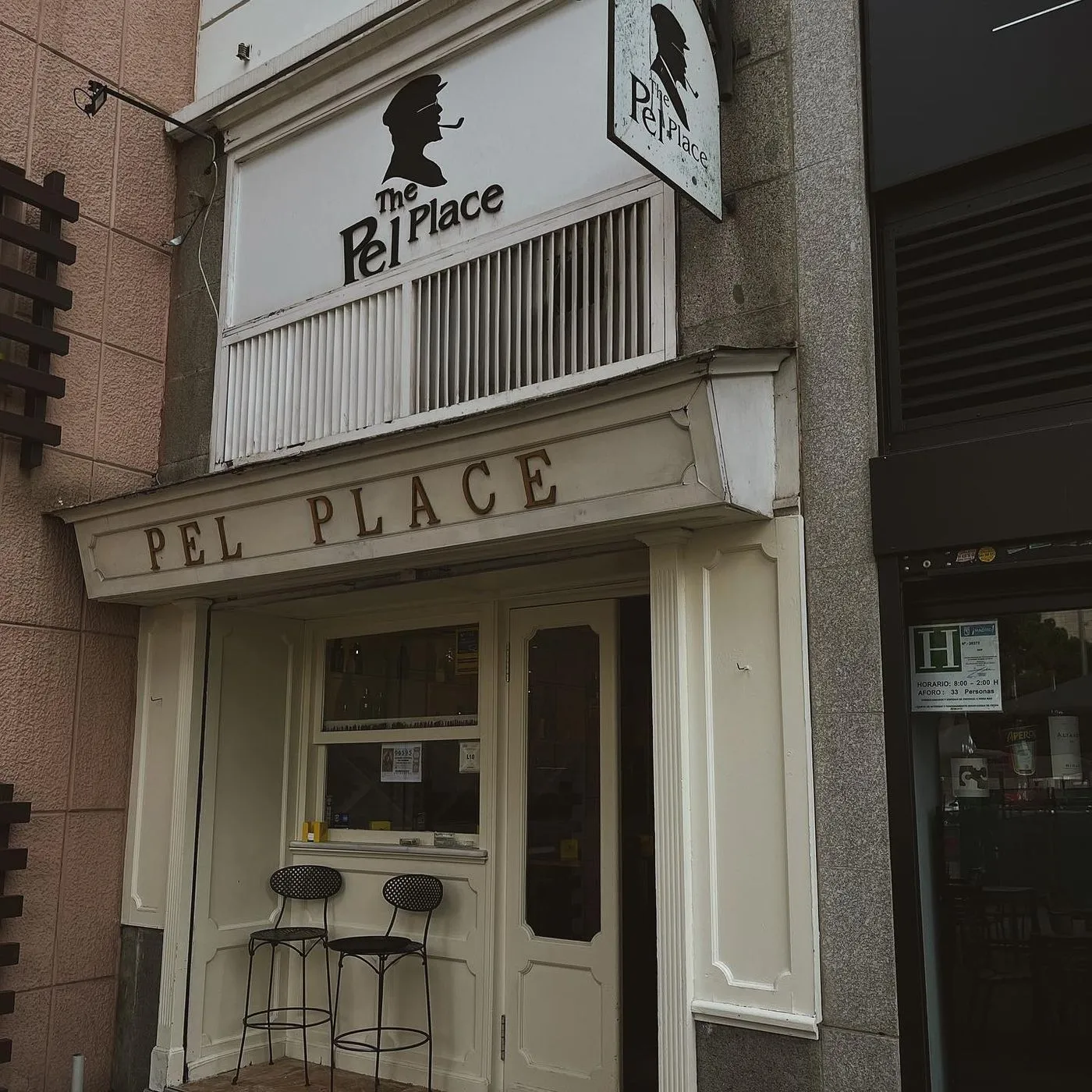 Fachada blanca con el rótulo del hombre de gorra y pipa, las letras doradas «PEL PLACE» y dos taburetes