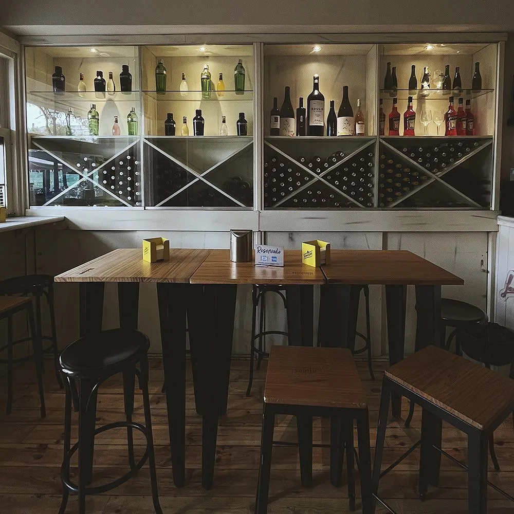 Mesas altas de madera con taburetes frente a una vitrina de botellas